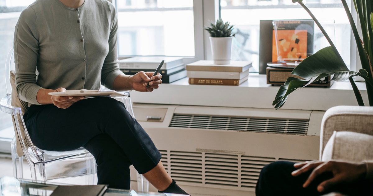 A counsellor sitting with a notebook, listening to a client in a quiet room with plants and daylight.