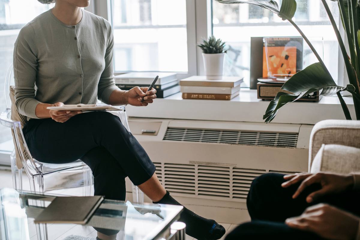 A counsellor sitting and listening to someone in a quiet, plainly furnished room with daylight and a plant.